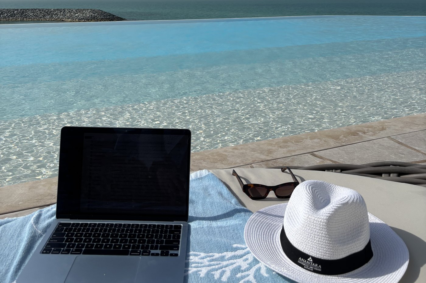 A laptop, sunglasses and a sun hat on a towel beside an infinity pool